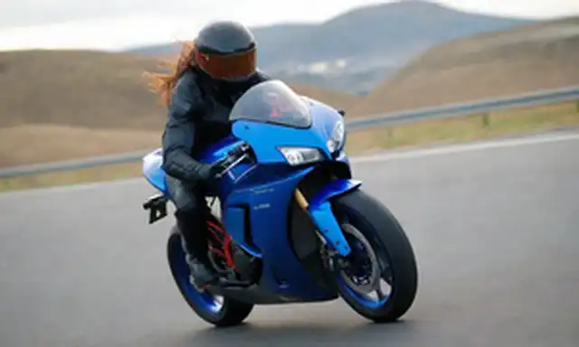 Una bici elegante y futurista con un brillante esquema de color azul, líneas curvas y acentos rojos brillantes; un jinete en movimiento, con pelo barrido por el viento y una gran sonrisa, volando por un camino escénico o carretera; un fondo borroso de paisaje urbano o campo, transportando velocidad y libertad