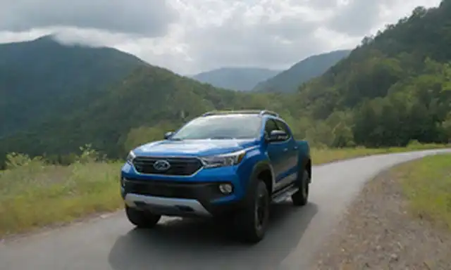Camión de camioneta 4x4 con puerto de carga eléctrico a lado, faros futuristas y logotipo de BYD en la parrilla; camión que conduce en carretera de montaña de viento, rodeado de vegetación y cielos azules