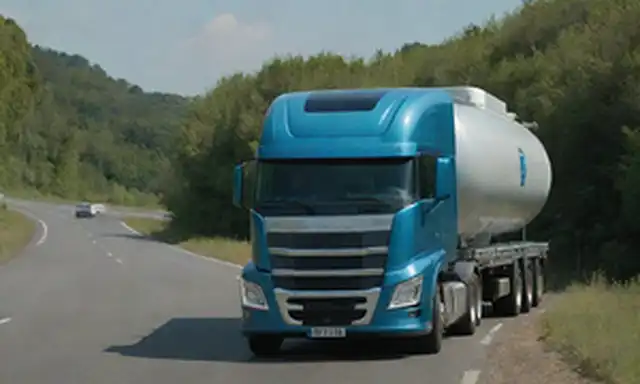 Un camión futurista en una carretera, rodeado de vegetación, con un fondo azul del cielo, tanques de gas de hidrógeno visibles a ambos lados, conductor en cabina, panel de control de ruta y nivel de combustible, señales de transporte sostenible