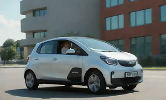 5 puertas con diseño elegante, vehículo eléctrico con ruedas futuristas, tamaño compacto para la conducción de la ciudad, amplio interior con dos asientos y pequeño tronco, pareja sonriente disfrutando de un paseo juntos, paisaje urbano en el fondo, día soleado con cielo azul