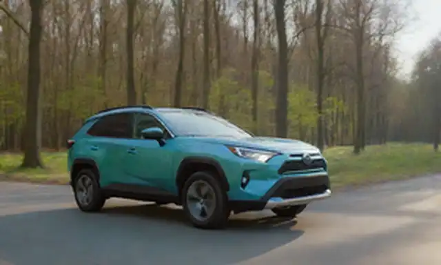2023 Toyota RAV4 Coche híbrido que conduce en un paisaje verde, rodeado de árboles y un cielo azul, con un toque de sol, mostrando su diseño elegante, faros LED, e insignia híbrida en el lado, contra un fondo borroso de enfoque suave