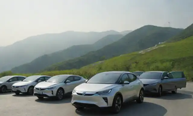 2025 Coches eléctricos chinos alineados en una carretera montañosa, luz solar brillante, colinas verdes en el fondo, diseño futurista elegante, estaciones de carga cercanas, peatones y coches que se mueven libremente, turbinas de viento o paneles solares visibles en las azoteas, paisaje urbano que se desvanecen en la distancia