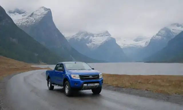 Montañas escandinavas, fiordo sereno, camioneta azul eléctrica conduciendo a través de paisajes maliciosos, bandera de Noruega en el fondo, terreno accidentado, picos cubiertos de nieve, espectacular paisaje nublado