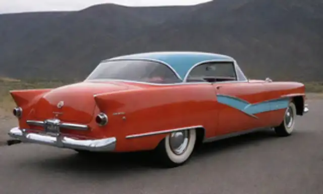 Carro de estilo 1950 con elementos de diseño futuristas de inspiración nuclear, tales como tubos azules brillantes, símbolos atómicos, y una forma elegante y aerodinámica; cuerpo metálico con un esquema de color rojo audaz; aletas estilizadas en la espalda; y una parrilla prominente con un toque de detalles tipo cohete