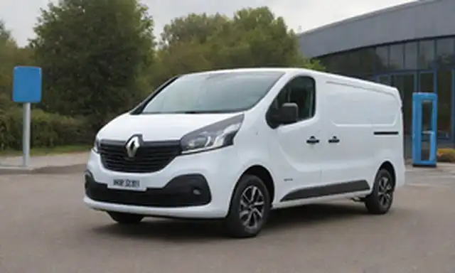 Blanco Renault Trafic Furgón E-Tech Electric van en un estacionamiento, con puertas traseras abiertas y cable de carga enchufado en el puerto de vehículos, luz solar brillante iluminando su elegante corte azul eléctrico, estacionado junto a una estación de carga con luces LED verdes, transportando modernidad y eco-amigración