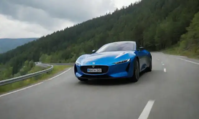 Eléctrico coche azul con diseño elegante, conduciendo en carretera de montaña con fondo de bosque verde, sol brillando brillantemente sobre la cabeza, nubes que se desvían perezosamente a través del cielo, sombras sutiles que resaltan líneas curvas de vehículo, estado de ánimo global es vibrante y ecológica, sutil arquitectura nórdica insinúa en la distancia