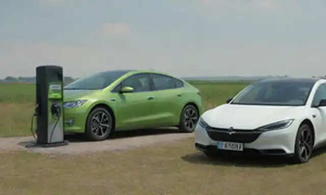 2 coches, campos verdes, día soleado, estación de carga eléctrica en el fondo, logotipos de vehículos híbridos en las puertas laterales