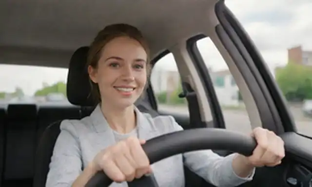 Vehículo eléctrico, fondo blanco, entorno urbano, conducción de autos, persona sentada en asiento de conductores, sonriendo, mirando adelante, manos en volante, postura relajada, paisaje urbano borroso en el fondo, soleado día con cielo azul y nubes suaves