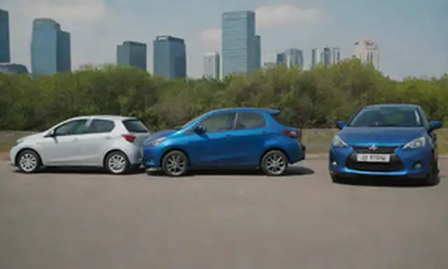 Tortillas de 3 puertas, aparcadas lado a lado, Toyota Yaris en azul, Mazda2 híbrido en plata, vista frontal, con carreteras y edificios borrosos en fondo, luz solar de la tarde sombras de fundición