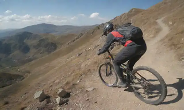 bicicleta de montaña en acción, jinete inclinado hacia adelante, terreno robusto, camino rocoso, fondo escénico, cielo soleado, ruedas borrosas, casco de jinetes, guantes y chaqueta visible