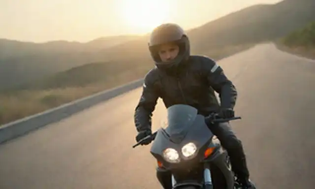 Motocicleta de 2 ruedas, jinete con casco y engranajes protectores, carretera o paisaje montañoso, puesta del sol en el fondo, cabello barrido por el viento, ligero borrón del movimiento, sentido de libertad y aventura