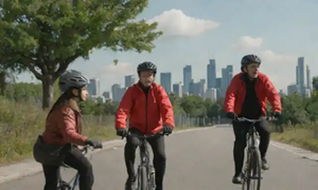 2-4 personas en bicicletas, una en chaqueta roja, otra sujetando un casco, contra un cielo azul con pocas nubes, con un paisaje urbano en el fondo, ciclistas interactuando, carreteras y edificios borrosos, luz solar brillante, vegetación, arquitectura moderna