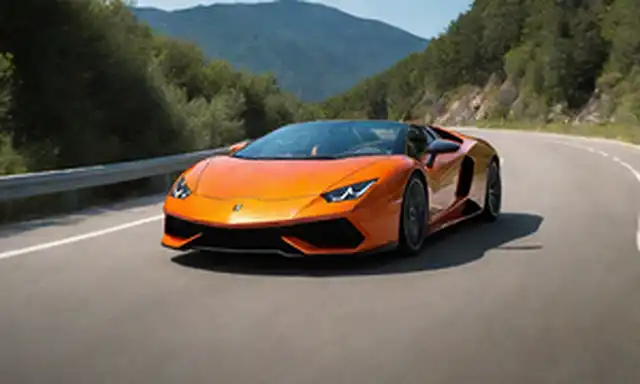 2023 Lamborghini Revuello coche deportivo híbrido en naranja metálica con puertas tijeras abiertas, mostrando interior elegante y avanzado sistema de infotenimiento, junto con un camino de montaña enrollador, luz solar brillante y árboles de fondo borroso, capturando su poderosa postura, diseño aerodinámico y movimiento dinámico