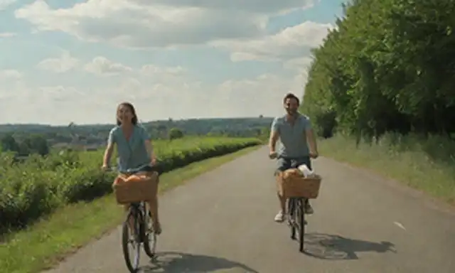 2 personas, cabalgando en una carretera, soleado día, sonriendo, manos en manillar, canastas en la espalda, paisaje urbano, caminos de bicicleta, vegetación, nubes, cielo azul