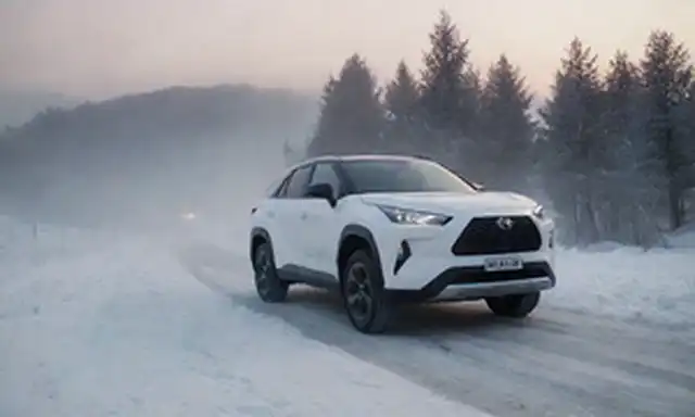 carretera nevada, Toyota bZ4X SUV eléctrico en foco, paisaje de invierno con árboles y colinas, ligeramente iluminado, niebla helada, faros brillantes, suave pendiente, ambiente sereno